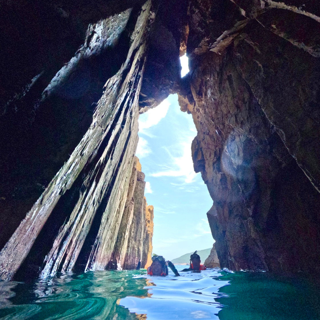 Two people in Hong Kong Geopark exploring a large rocky cave entrance with clear blue water.